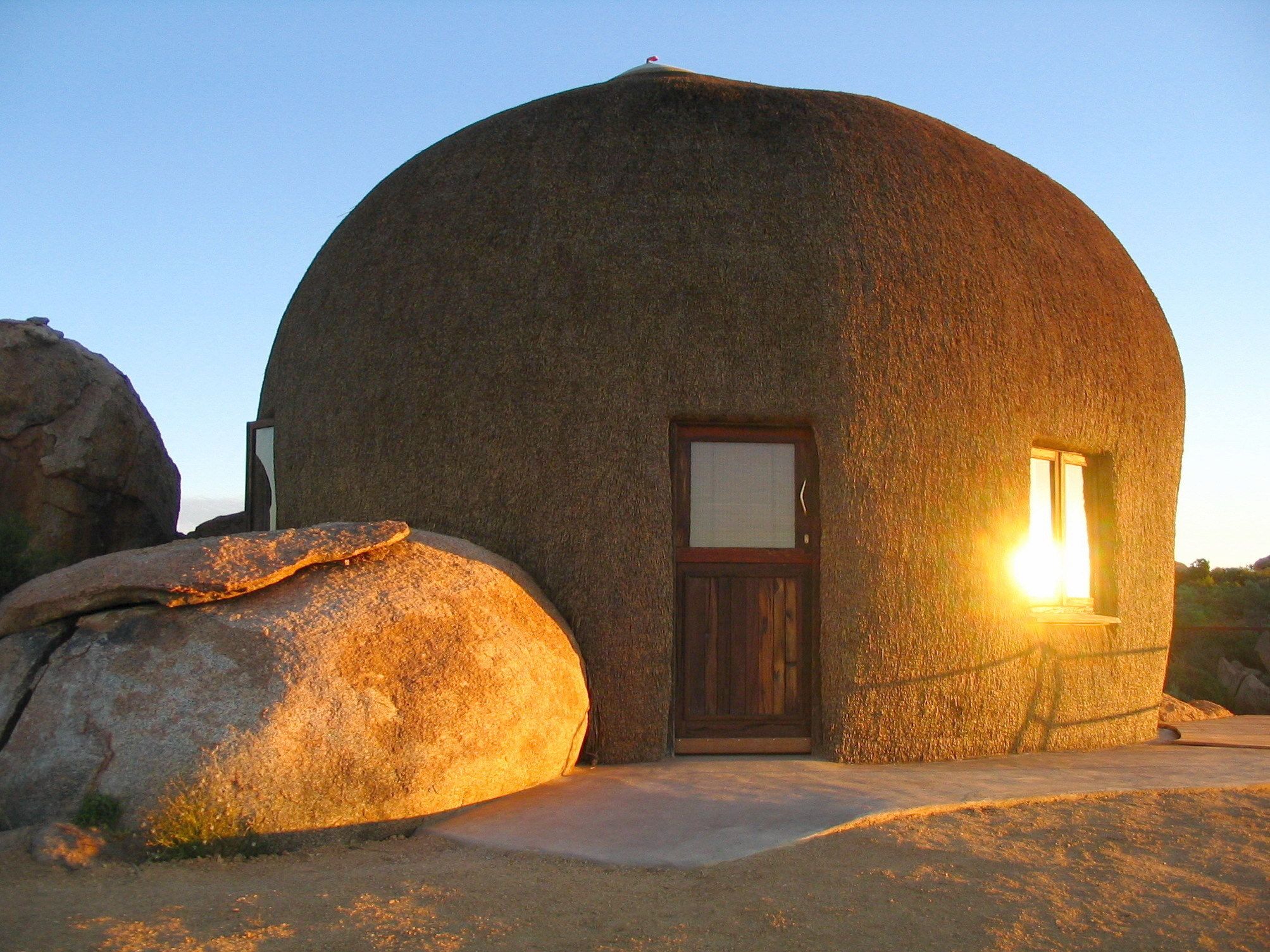 Un bâtiment sphérique en pierre, doté d’une porte et d’une fenêtre en bois, entouré de rochers.