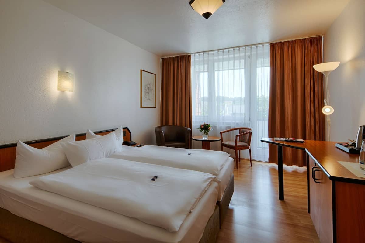 Een hotelkamer met twee bedden, een bureau, een stoel, een klein tafeltje met een vaas, en een raam met gordijnen.