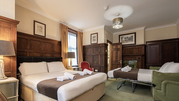 A hotel room with a large bed, a smaller bed, a green armchair, a wooden cabinet, and a fireplace. - Barrington Lodge Hotel  (Crawley)