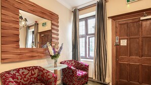 A hotel lobby with a wooden reception desk, a mirror, a red patterned armchair, a small table with a vase of flowers, and a large wooden door. - Barrington Lodge Hotel  (Crawley)