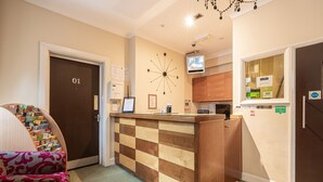 A hotel lobby with a reception desk, a check-in counter, a wall-mounted television, and a patterned armchair. - Barrington Lodge Hotel  (Crawley)