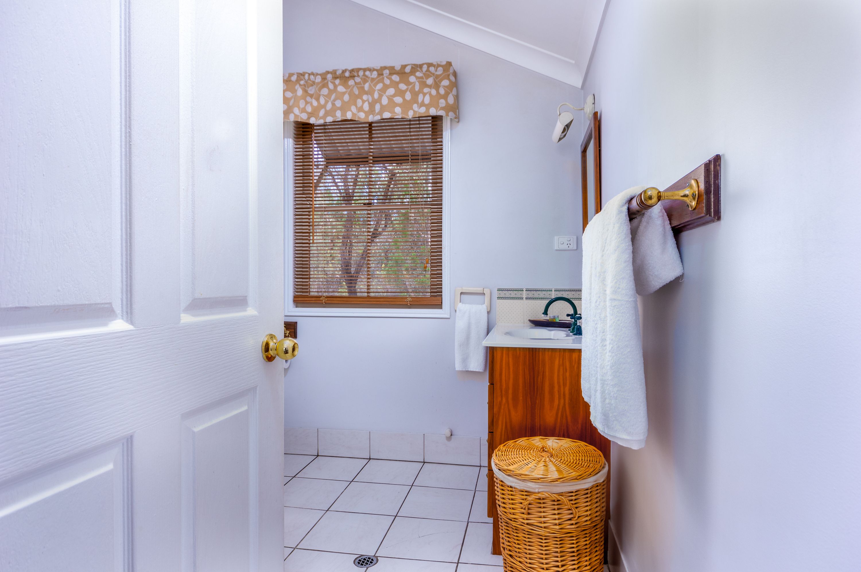 Un baño con una puerta blanca, una ventana con persianas, un lavamanos con una toalla y una cesta de mimbre.