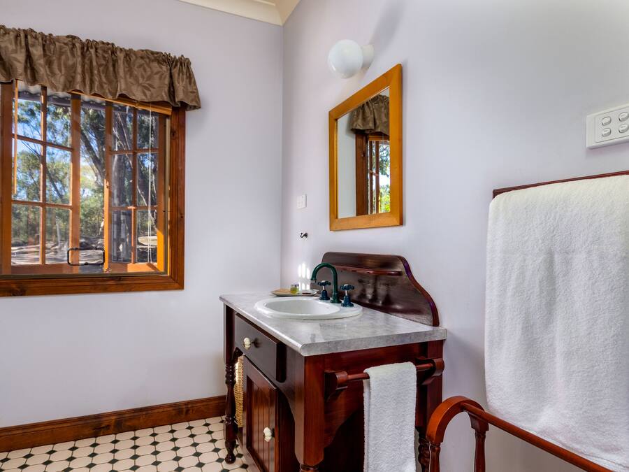 Un baño con tocador de madera, lavamanos blanco, espejo y ventana con vistas a árboles.
