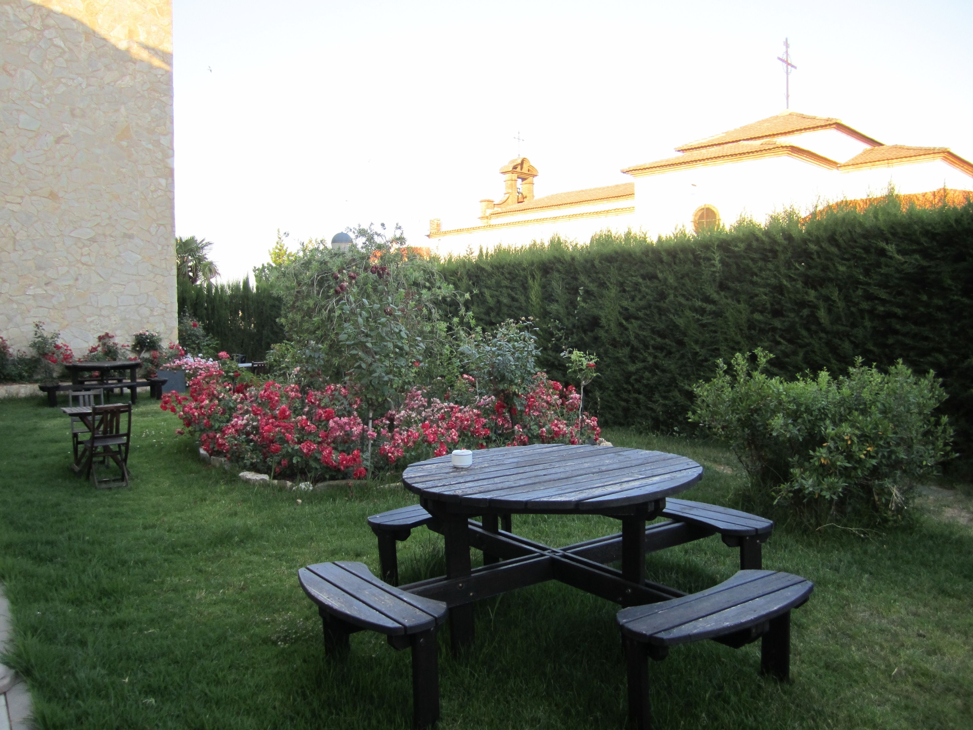 Eine Terrasse im Freien mit einem runden Tisch und Bänken, umgeben von einem gepflegten Garten mit verschiedenen Pflanzen und Blumen.