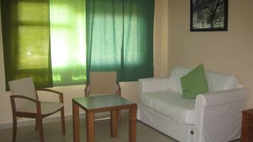 Una sala de estar con un sofá blanco, dos sillas de madera, una mesita y una ventana con cortinas verdes.