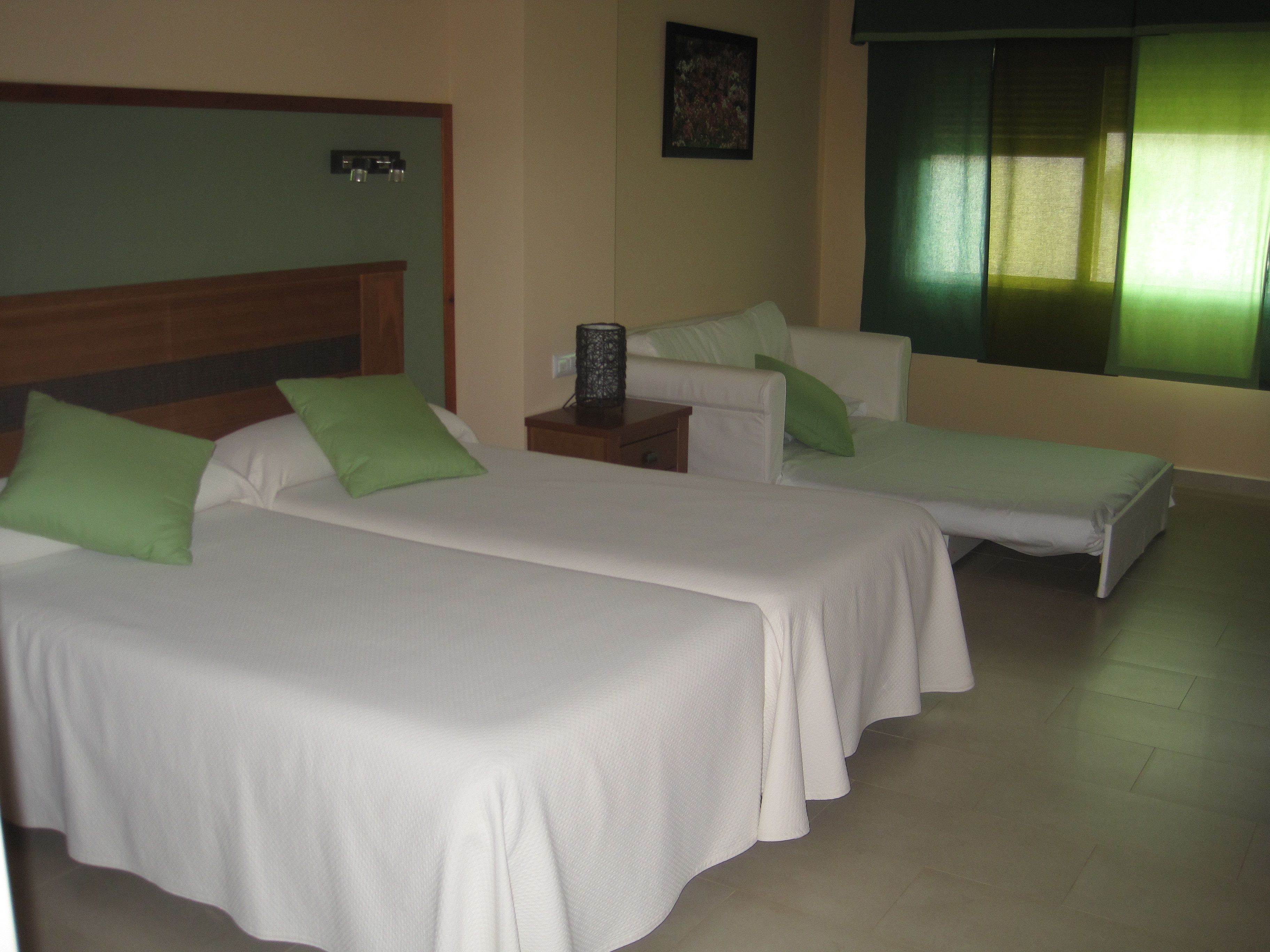 A hotel room with two single beds, each with a green pillow, a wooden headboard, a bedside table with a lamp, and a window with green curtains.