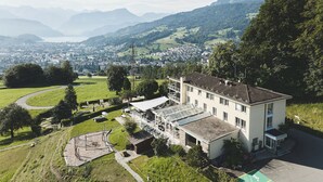 A modern building with a flat roof, large windows, and a balcony. It is surrounded by lush greenery and a winding road. - Hotel Sonnenberg (Kriens)