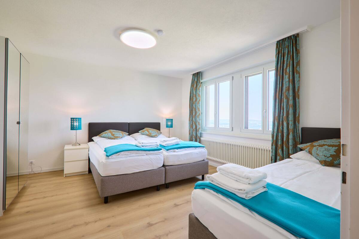 A hotel room with two beds, a nightstand, a lamp, a window with curtains, and a radiator.