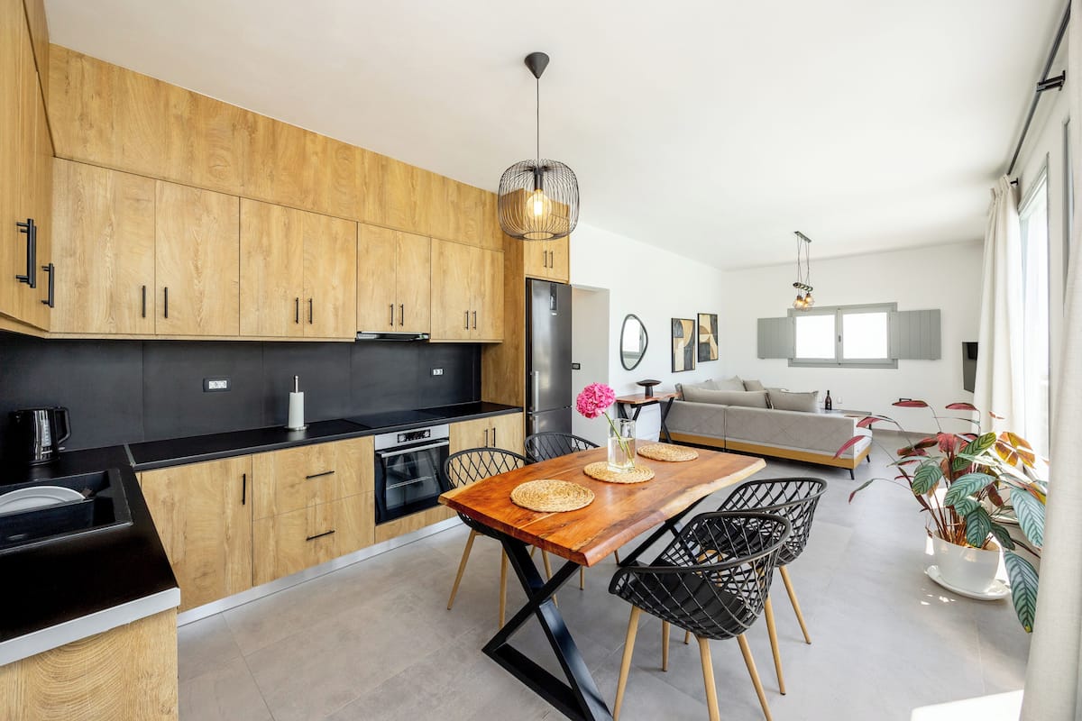 A modern kitchen with wooden cabinets, a black countertop, and a dining area with a wooden table and black chairs.