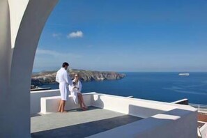 A couple on a balcony overlooking a coastal view with cliffs and the sea. - Thermes Luxury Villas by Mr and Mrs White (Santorini)