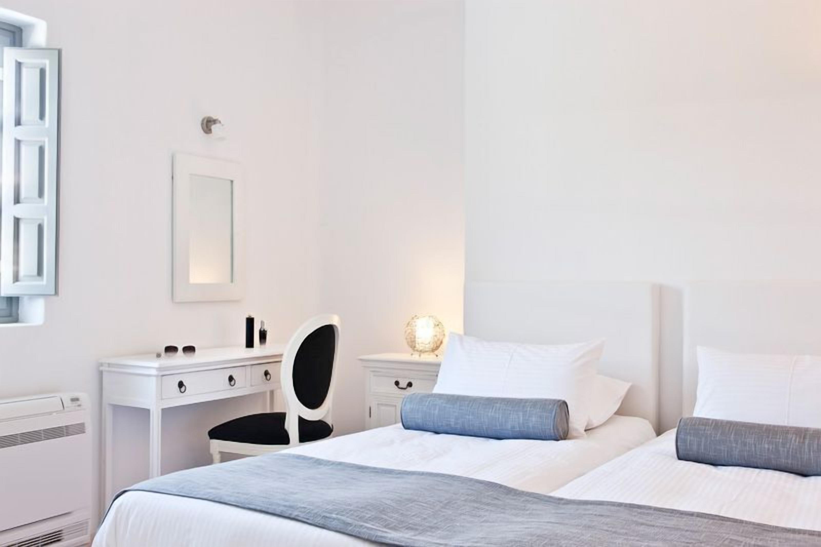 A hotel room with a bed, a desk, a chair, and a bedside table with a lamp.