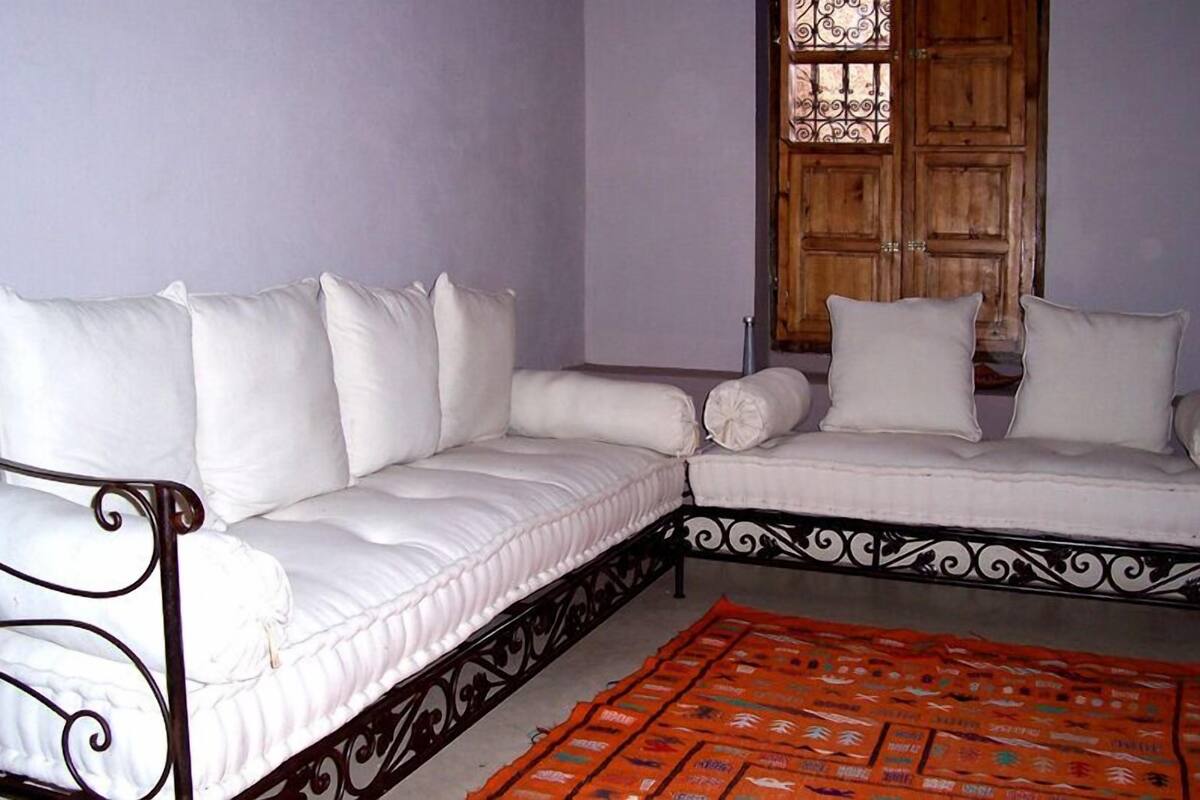 A room with a white sofa set against a light purple wall, a wooden door with decorative glass, and a patterned orange rug.