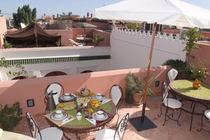 Eine Dachterrasse mit Esstisch, gedeckt für das Frühstück, umgeben von Stühlen und einem weißen Sonnenschirm, der Schatten spendet.