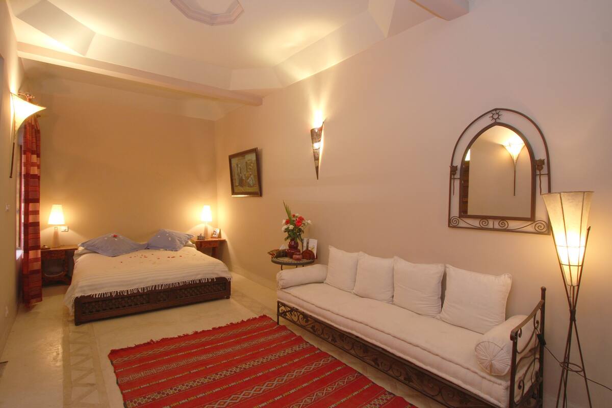 A bedroom with a bed, a sofa, a red patterned rug, a wall-mounted mirror, and two bedside lamps.