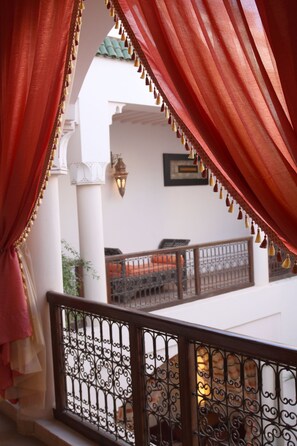 Ein Balkon mit aufwendig gestalteten Metallgeländern, roten Vorhängen und Blick auf ein Gebäude mit einem Ziegeldach und einer wandmontierten Leuchte.