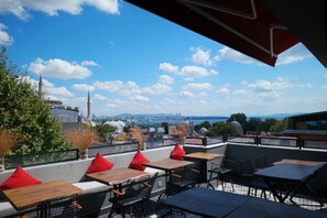 A rooftop terrace with tables and chairs, a view of a cityscape, and a clear sky.