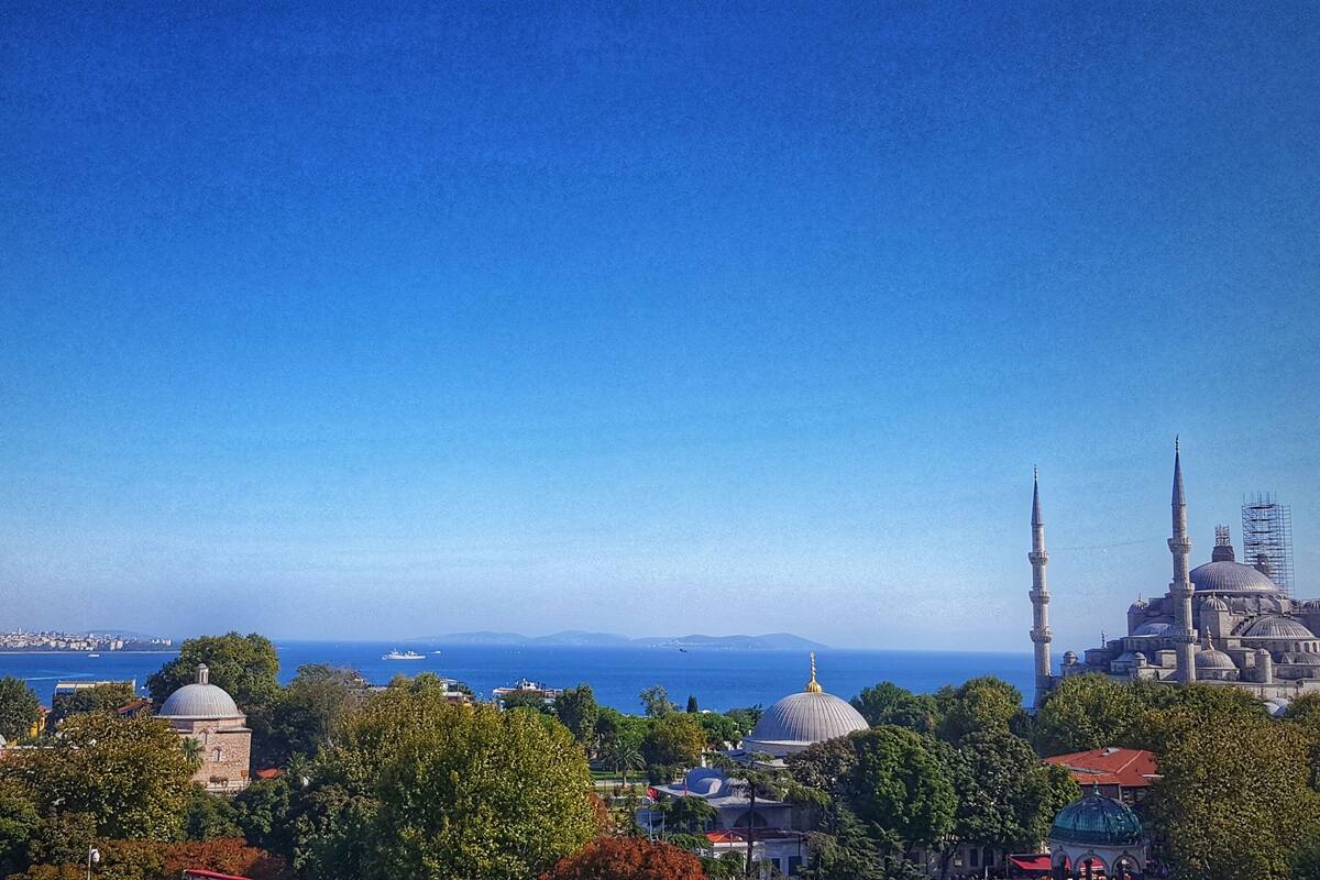 A cityscape with a prominent mosque featuring multiple minarets and domes, surrounded by lush greenery and a clear blue sky.