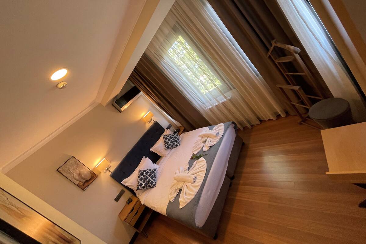 A hotel room with a bed, bedside table, lamp, and a view of the outdoors through sheer curtains.