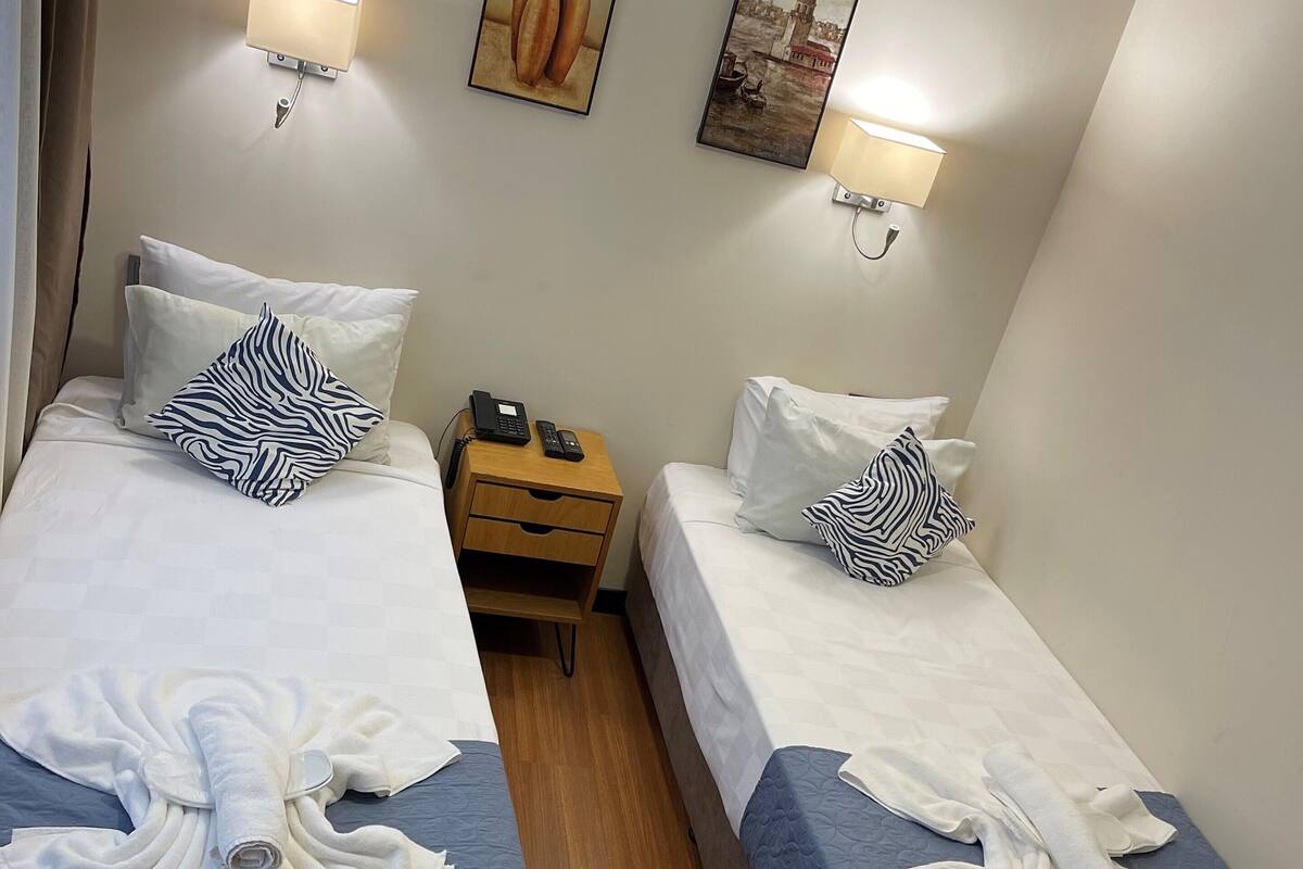 A hotel room with two beds, each with white linens and a towel folded on the bed. There are wall-mounted lamps, a nightstand with a phone, and artwork on the walls.