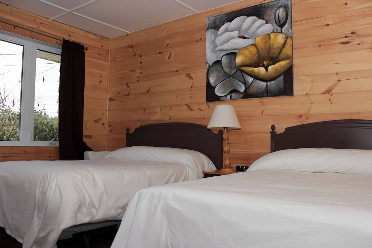A room with two beds, wooden walls, a painting, and a window with curtains.