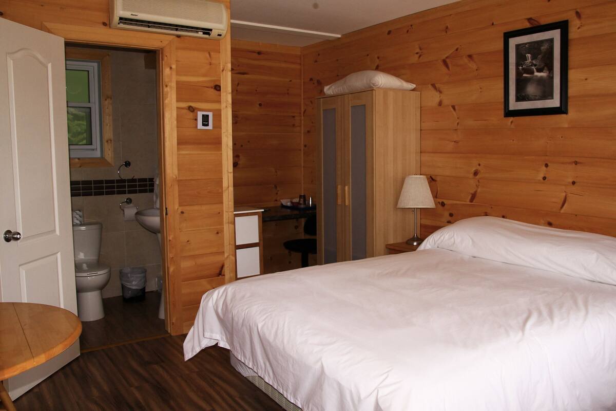 A wooden room with a bed, a bathroom, and a small desk.
