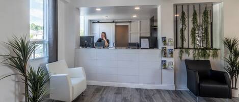 A reception area with a receptionist, a white reception desk, two chairs, and potted plants.