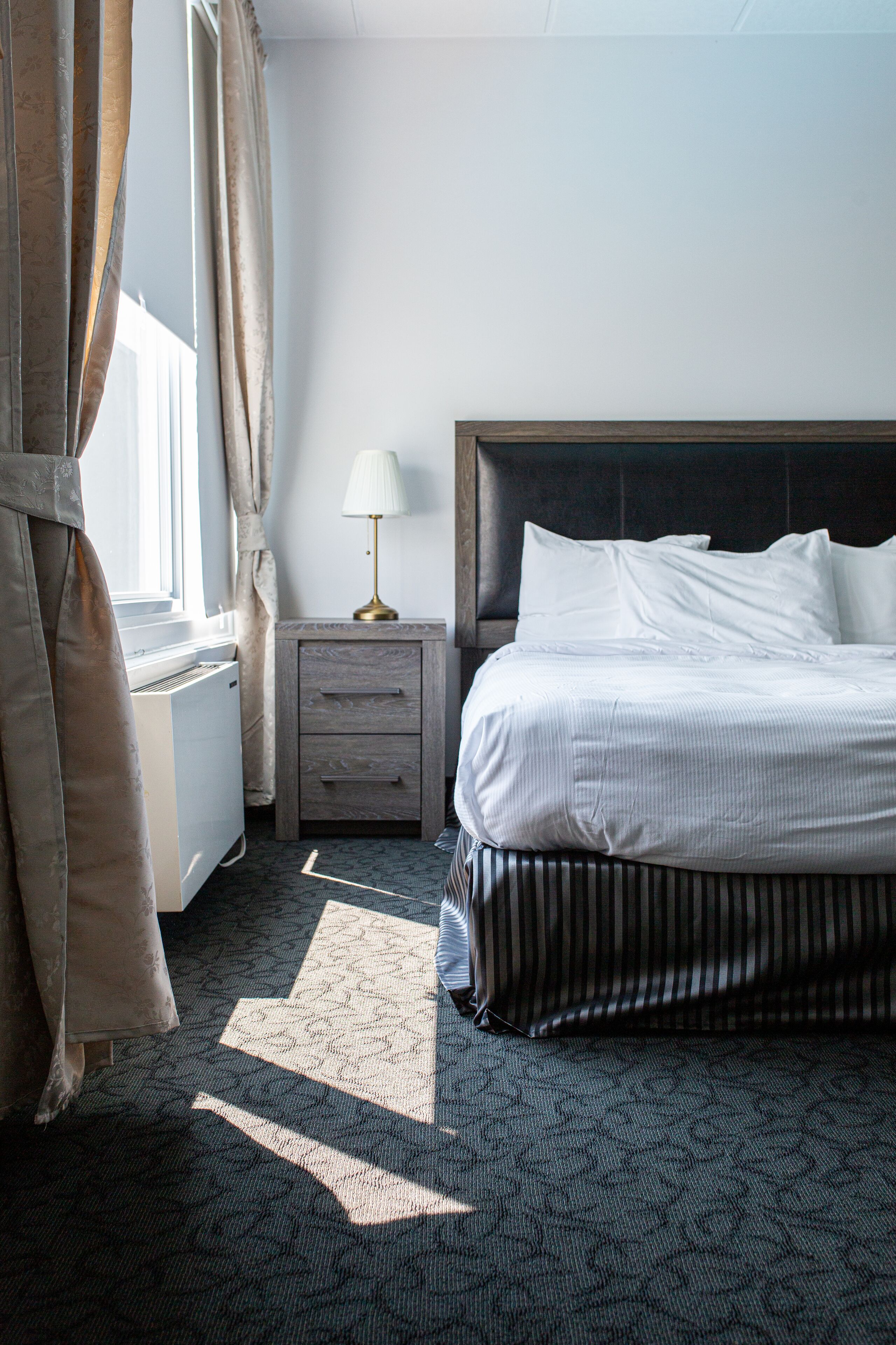 Een hotelkamer met een bed, nachtkastje, lamp en een raam met gordijnen.
