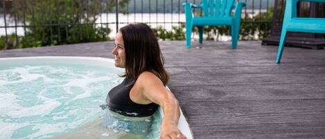 Una persona en un jacuzzi sobre una terraza de madera, con vista a un cuerpo de agua y un pabellón al fondo.