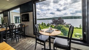 Un comedor con una mesa redonda y sillas, ventanas amplias con vistas a un lago y vegetación, y un televisor de pantalla plana montado en la pared.