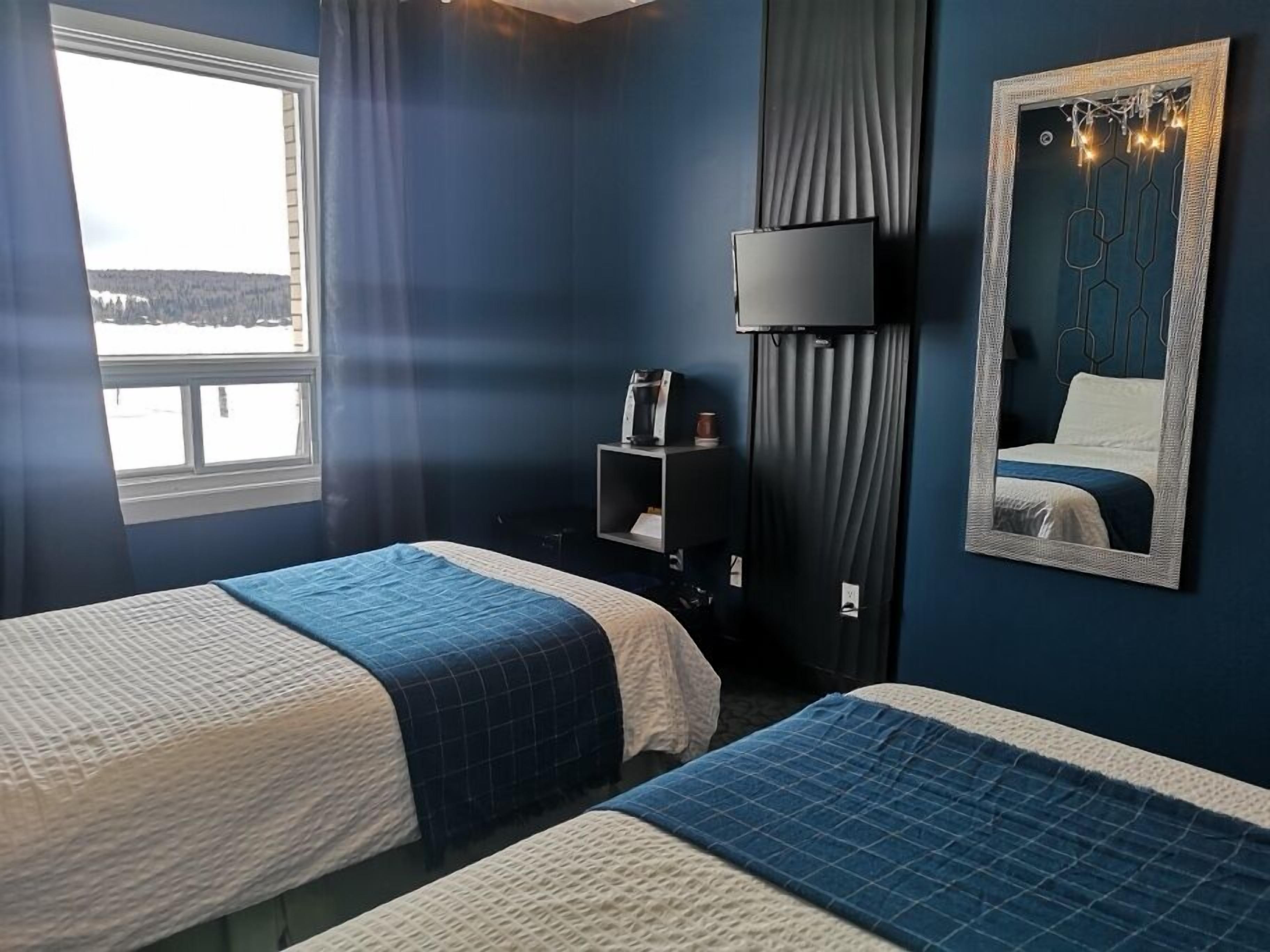A hotel room with two beds, a large window, a wall-mounted TV, and a mirror reflecting the bed.