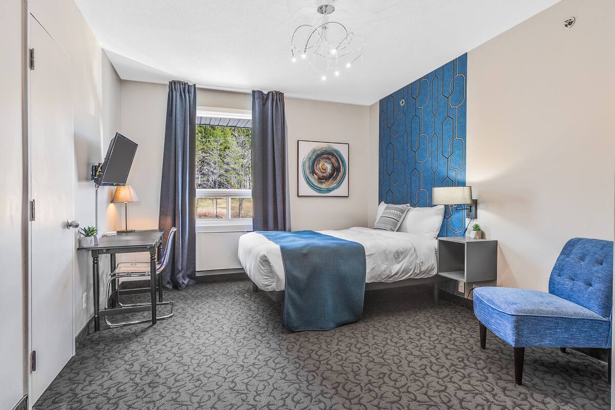 Una habitación de hotel con cama, escritorio con lámpara, silla, televisor y ventana con cortinas.