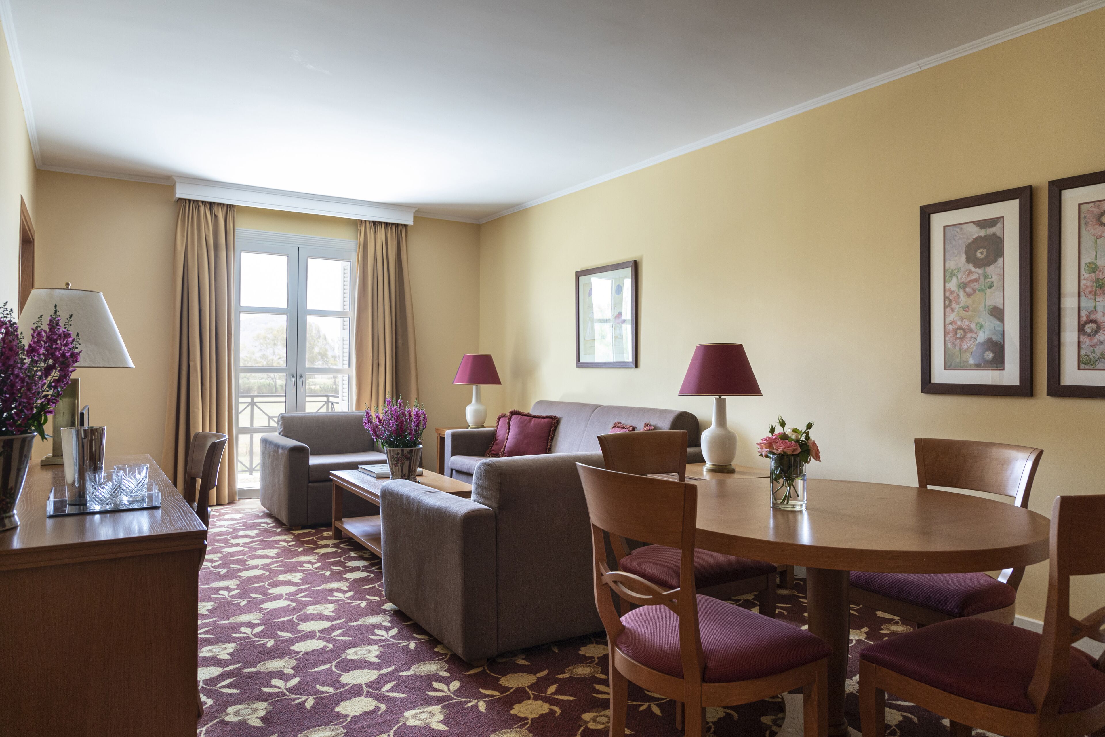 A hotel room with a sofa, armchair, dining table, chairs, a desk, lamps, and framed artwork on the walls.