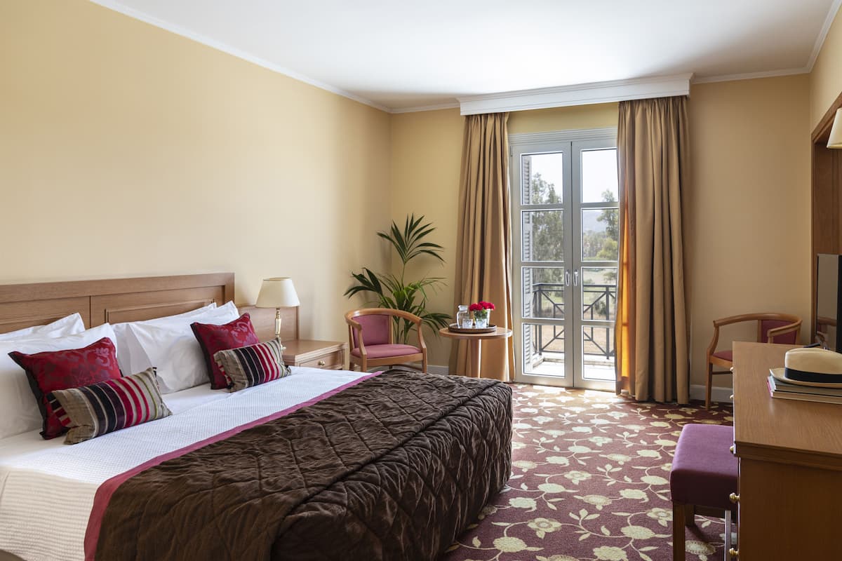 A hotel room with a bed, a desk, a chair, a small table with a plant, and a balcony with curtains.