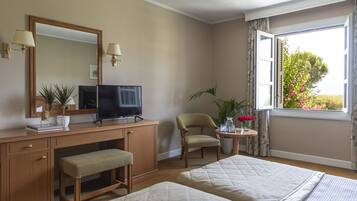 A hotel room with a wooden desk, a television, a chair, a bedside table with a vase of flowers, and two beds.