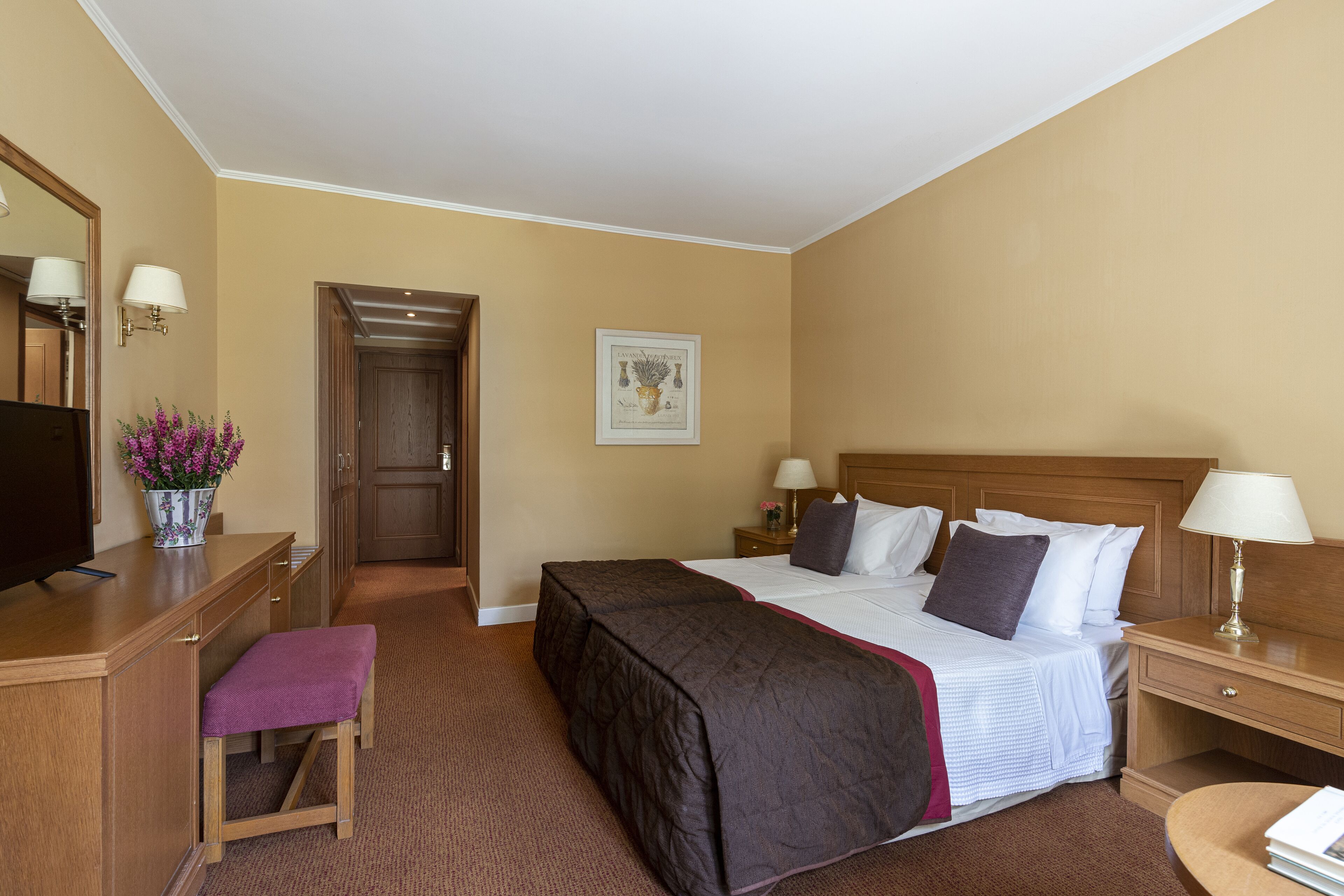 A hotel room with a bed, bedside tables, a desk with a chair, a television, and a door leading to another room.