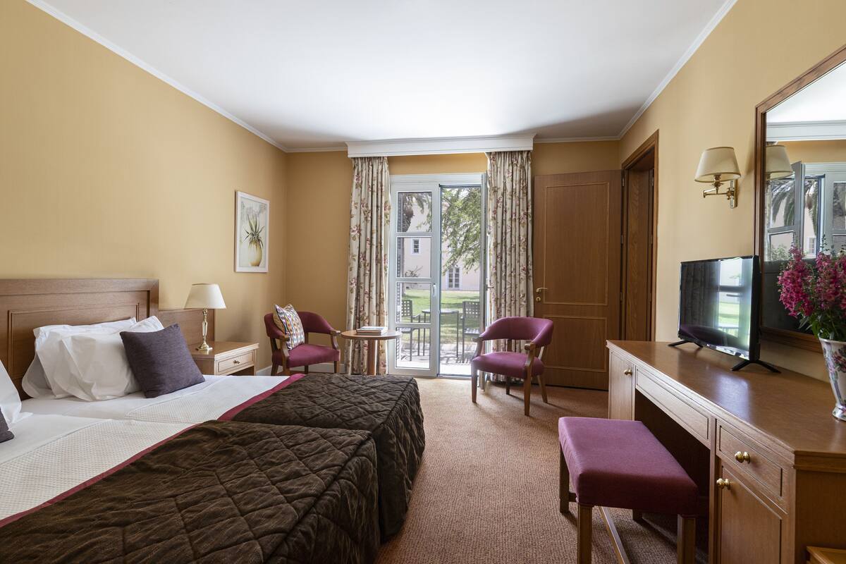 A hotel room with two beds, a desk, a chair, a TV, and a balcony with curtains.