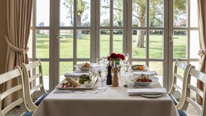 Mesa de comedor dispuesta con platos, vasos y un jarrón de flores rojas, con vistas a un jardín de césped a través de amplias ventanas.