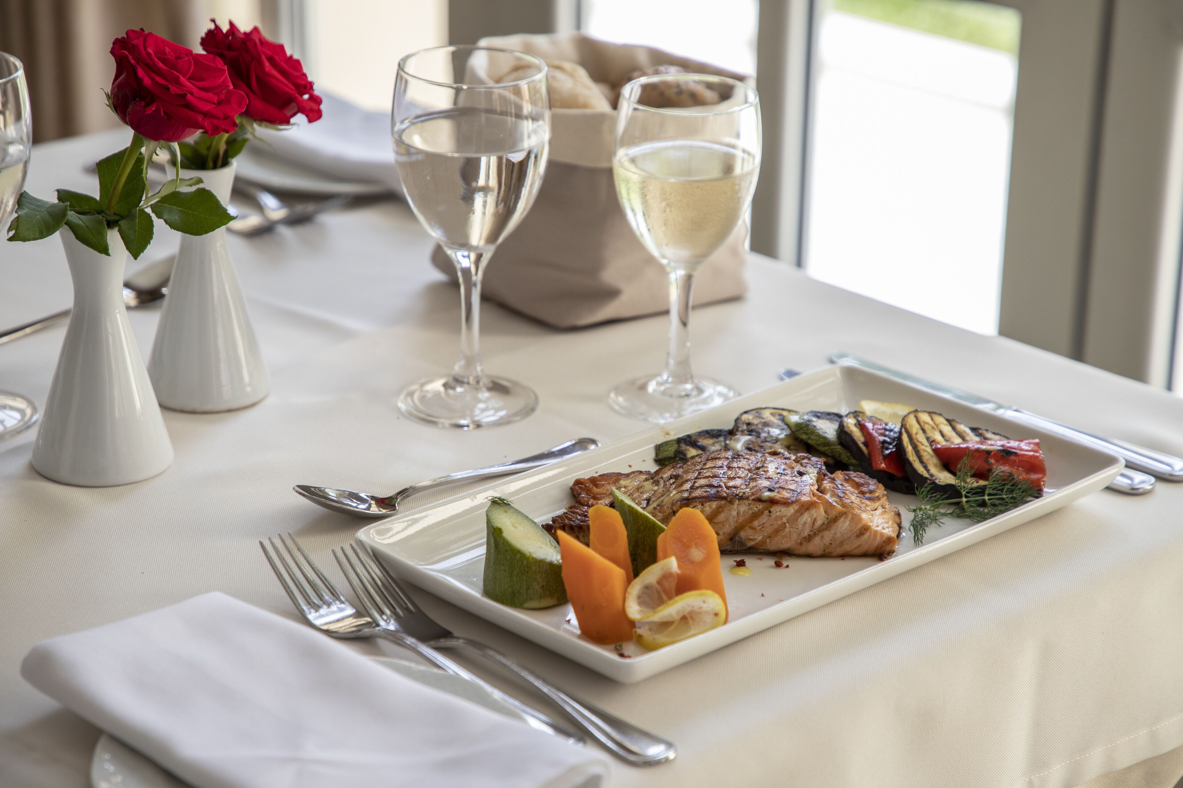 Ein Esstisch mit weißer Tischdecke, einem Teller mit Essen, zwei Vasen mit roten Rosen und zwei Gläsern Weißwein.