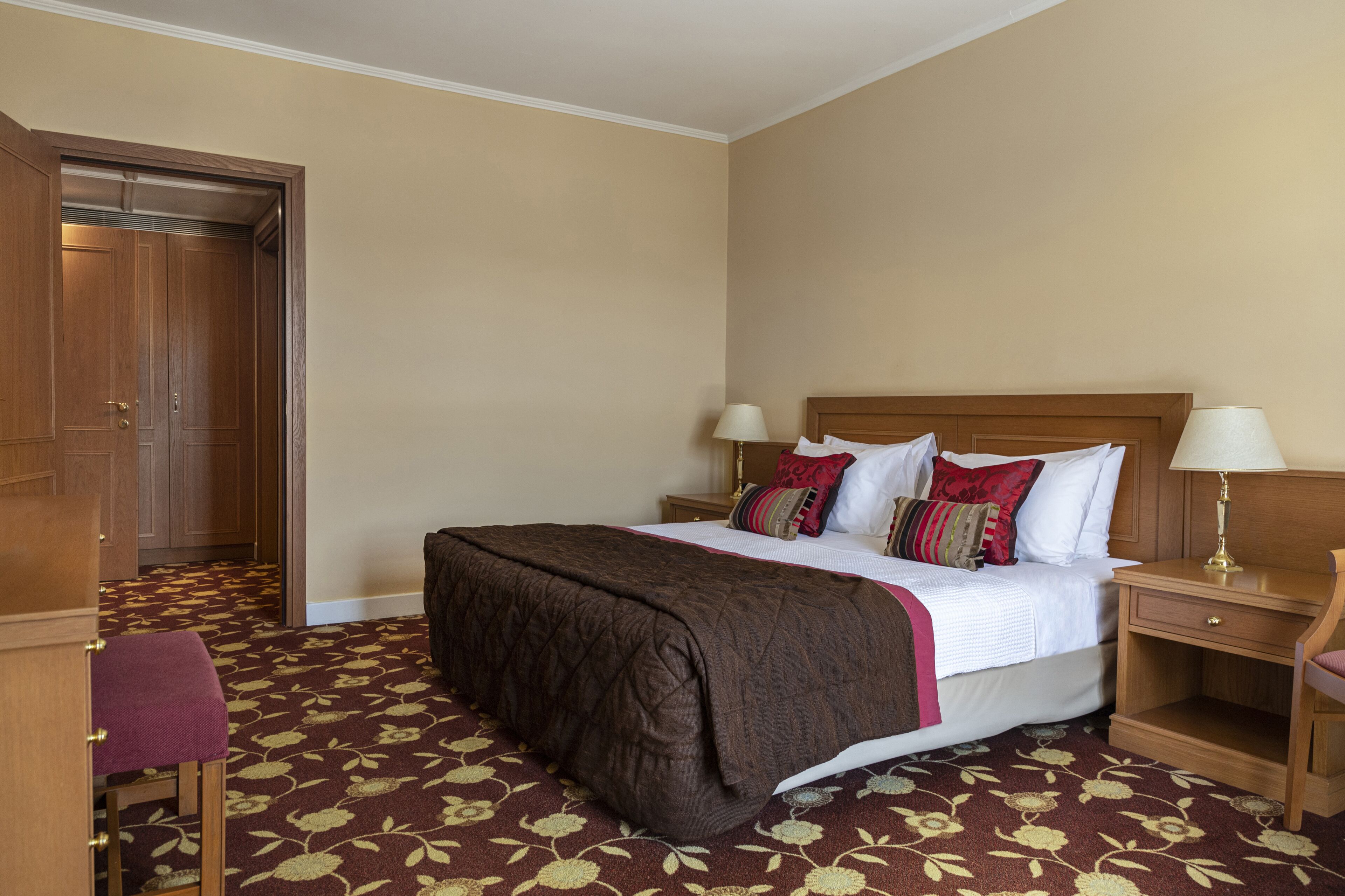 Habitación de hotel con una cama grande, cabecero de madera, dos mesitas de noche con lámparas, un armario de madera y suelo alfombrado.