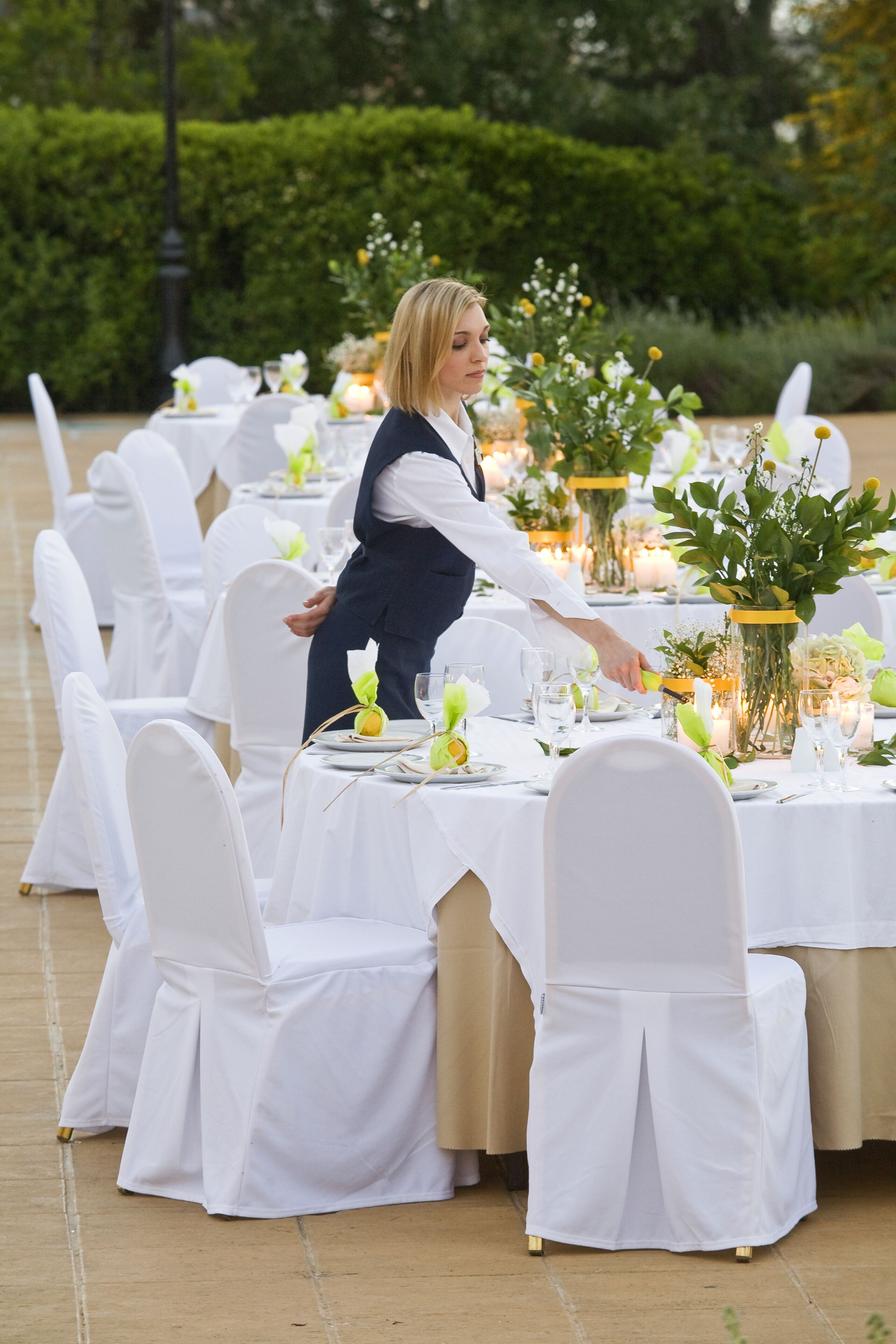 A banquet hall with round tables set for a formal event, surrounded by white chairs and greenery.