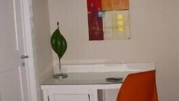 Ein kleines Zimmer mit einem weißen Schreibtisch, einem orangefarbenen Sessel, einem an der Wand montierten Fernseher und einem grünen Dekorationsobjekt auf dem Schreibtisch.