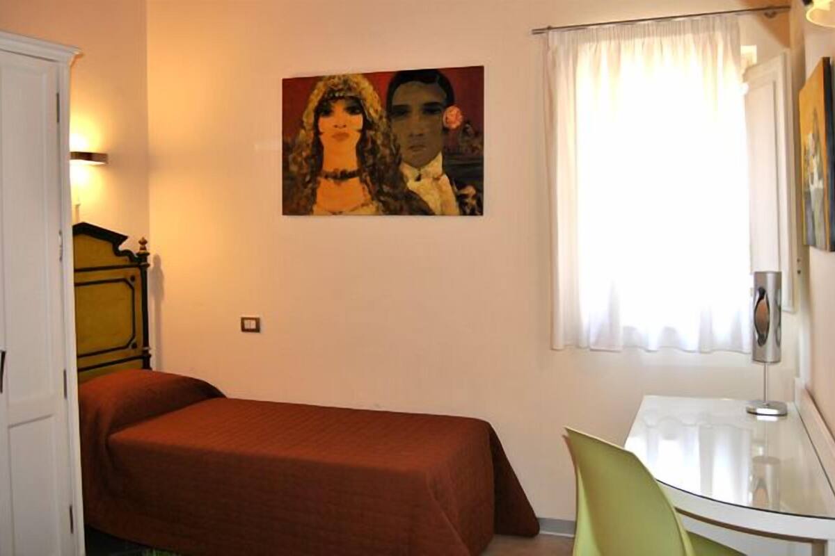 Une chambre à coucher avec un lit, un bureau, une chaise, un tableau et une fenêtre avec des rideaux.