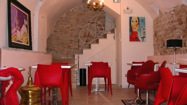 A dining area with red chairs and tables, a stone wall, a staircase, and a chandelier. - B&B Palazzo Ducale (Andria)