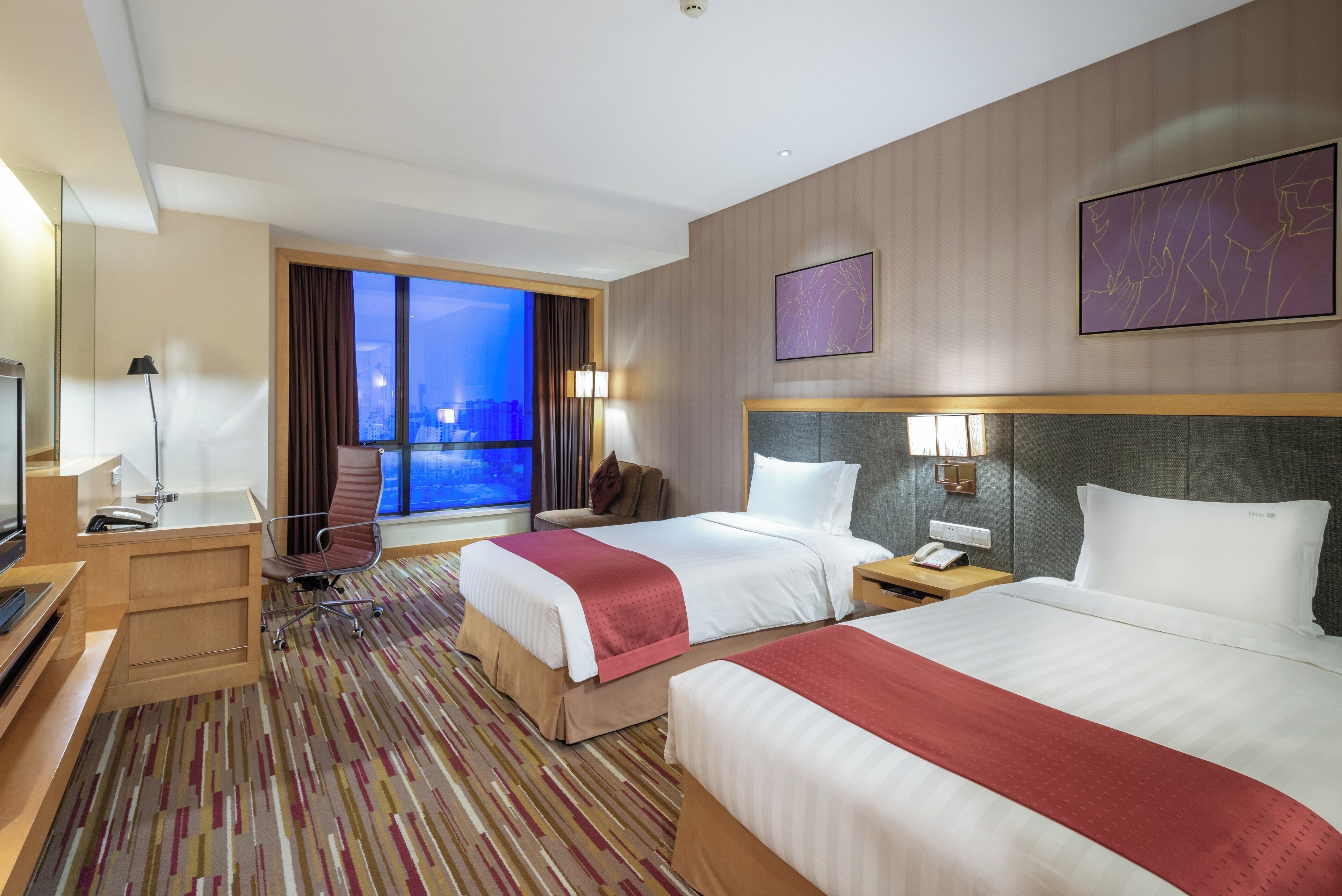 Een hotelkamer met twee bedden, een bureau, een stoel, een televisie en een raam met uitzicht op de stad.