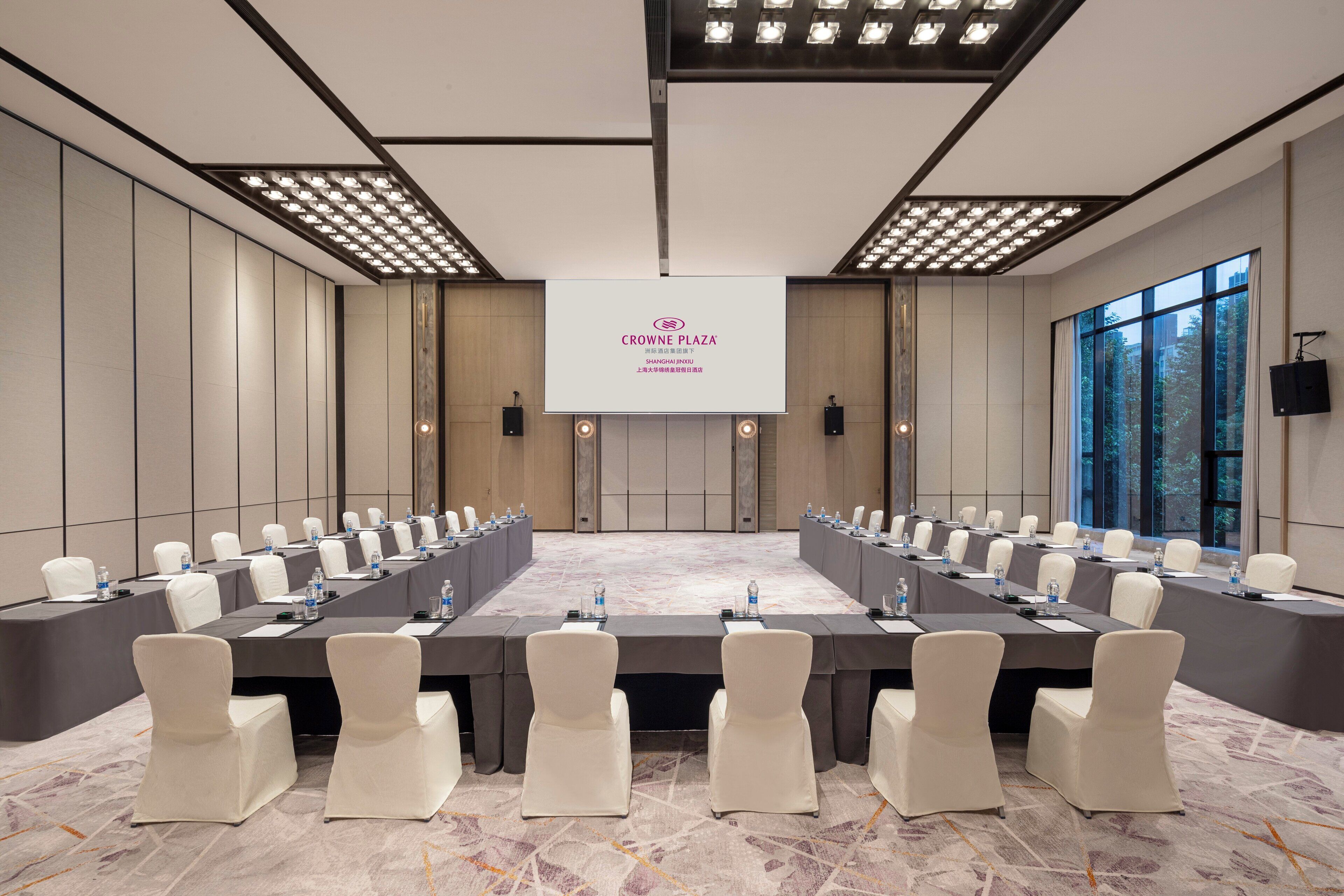 Ein Konferenzraum mit einem langen Tisch, weißen Stühlen und einem großen Bildschirm, der "CROWNE PLAZA SHANGHAI INN" zeigt.