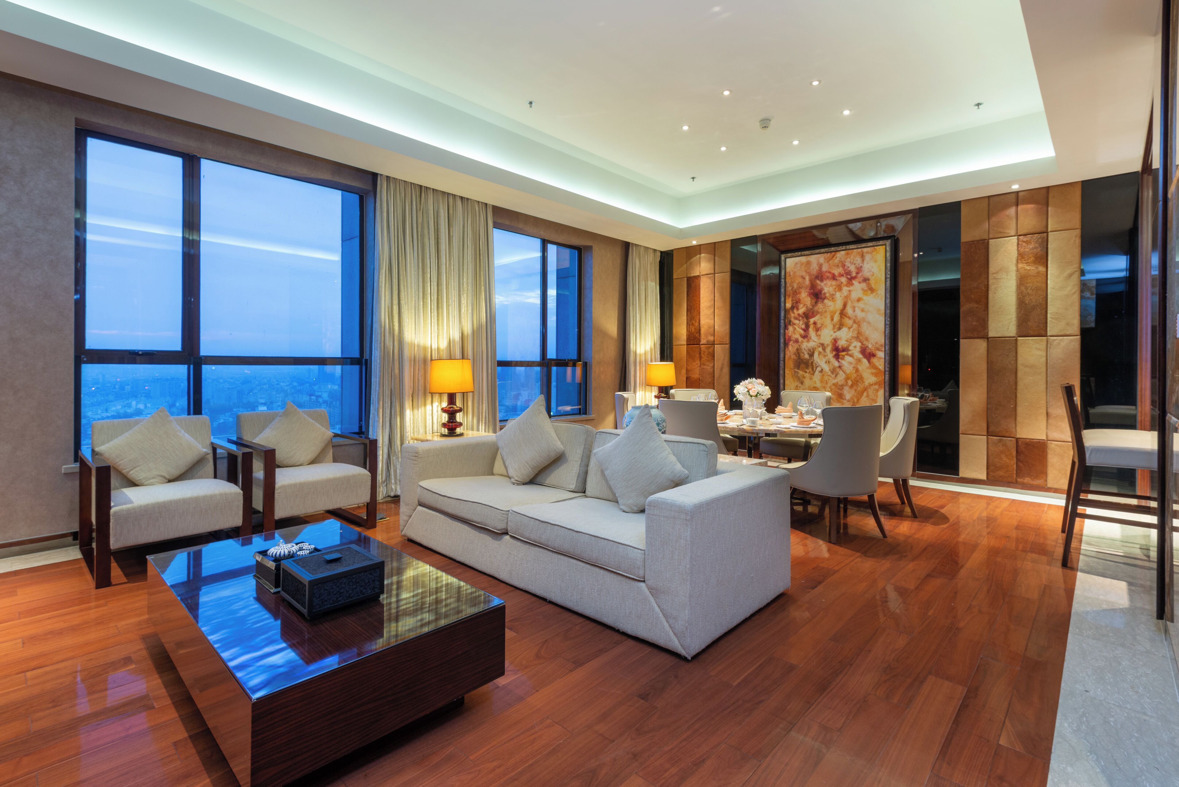 A modern hotel room with a sofa, chairs, a dining table, and a large window offering a city view.