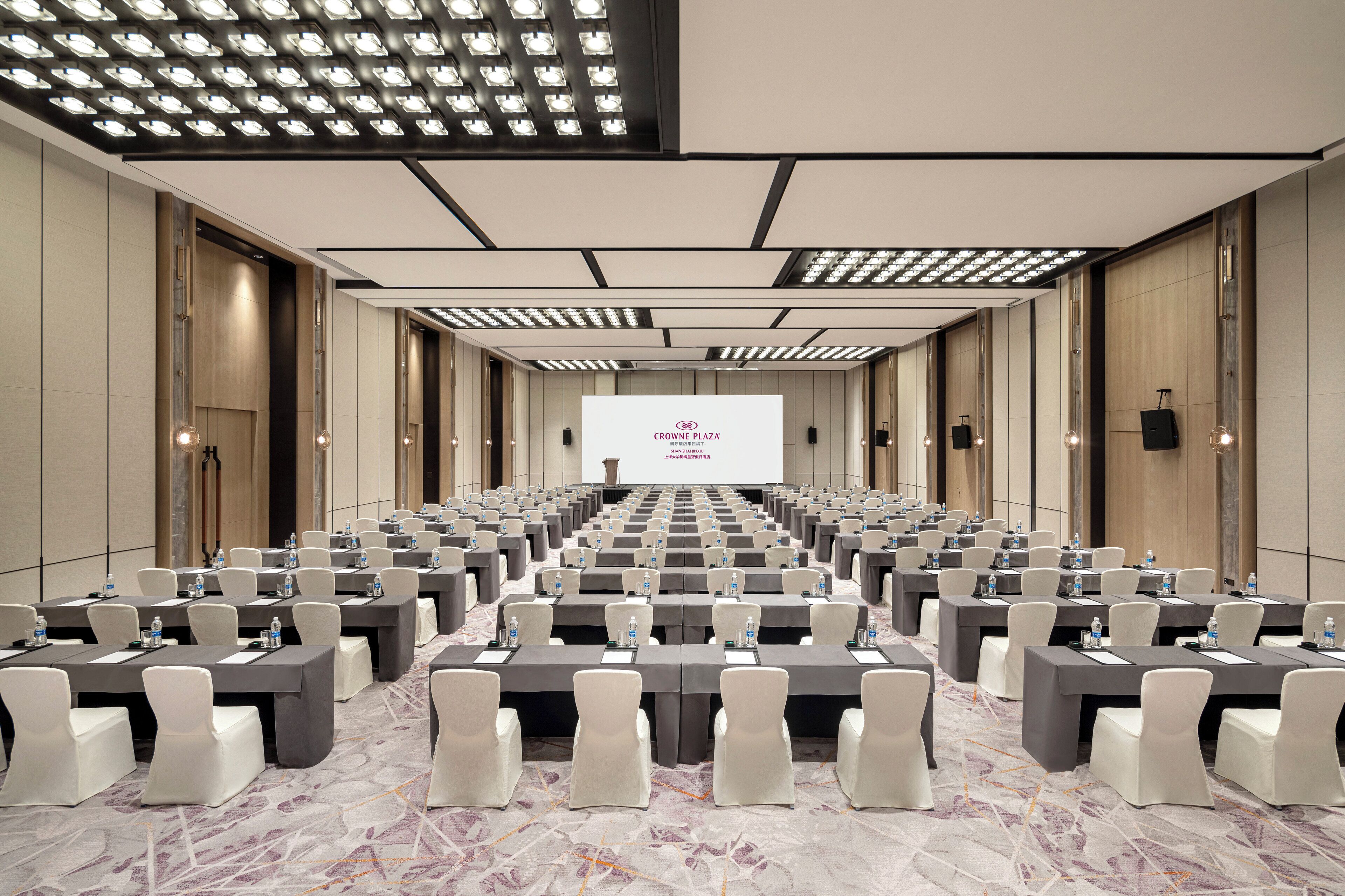 Une salle de conférence avec des rangées de tables et de chaises, un grand écran à l’avant et un design moderne au niveau du plafond.