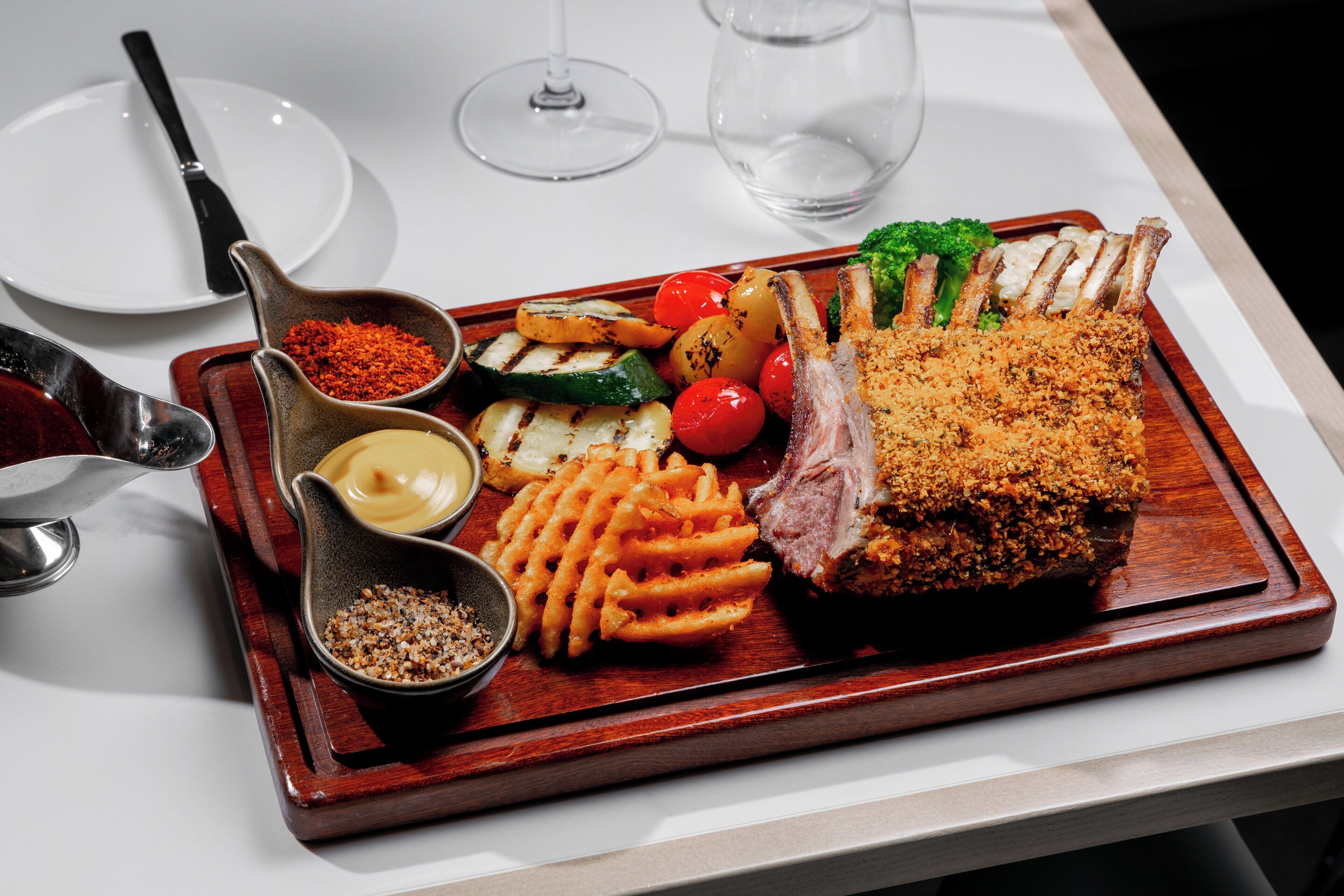 A wooden serving tray with a rack of lamb, grilled vegetables, and waffle fries on a table.
