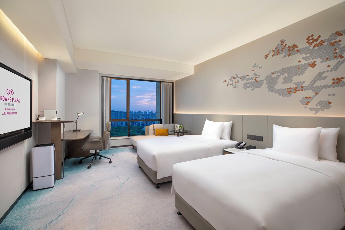 A hotel room with two beds, a desk, a chair, a small table, and a large window offering a city view.
