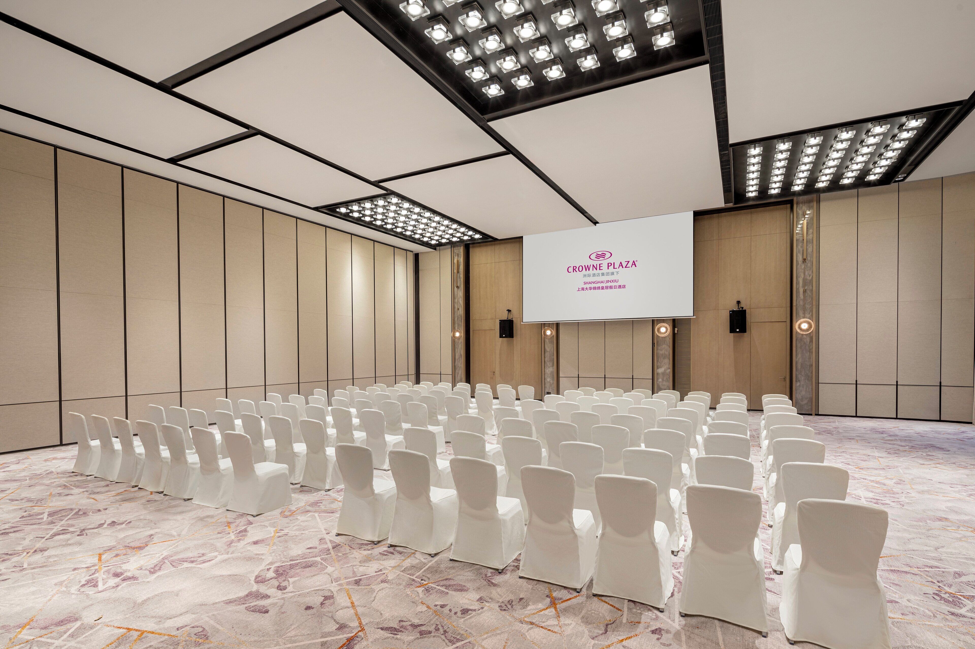 Un salón de conferencias con filas de sillas blancas, una pantalla de proyección y un piso de mármol.
